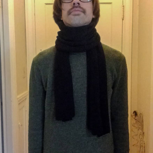 Person wearing a dark green sweater and black scarf in front of a wooden door.