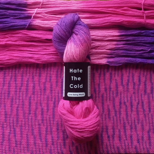 A skein of variegated Merino Wool yarn in pink and purple shades, wrapped with a black label that reads 'Hate The Cold', laid over a matching pink and purple knitted fabric.