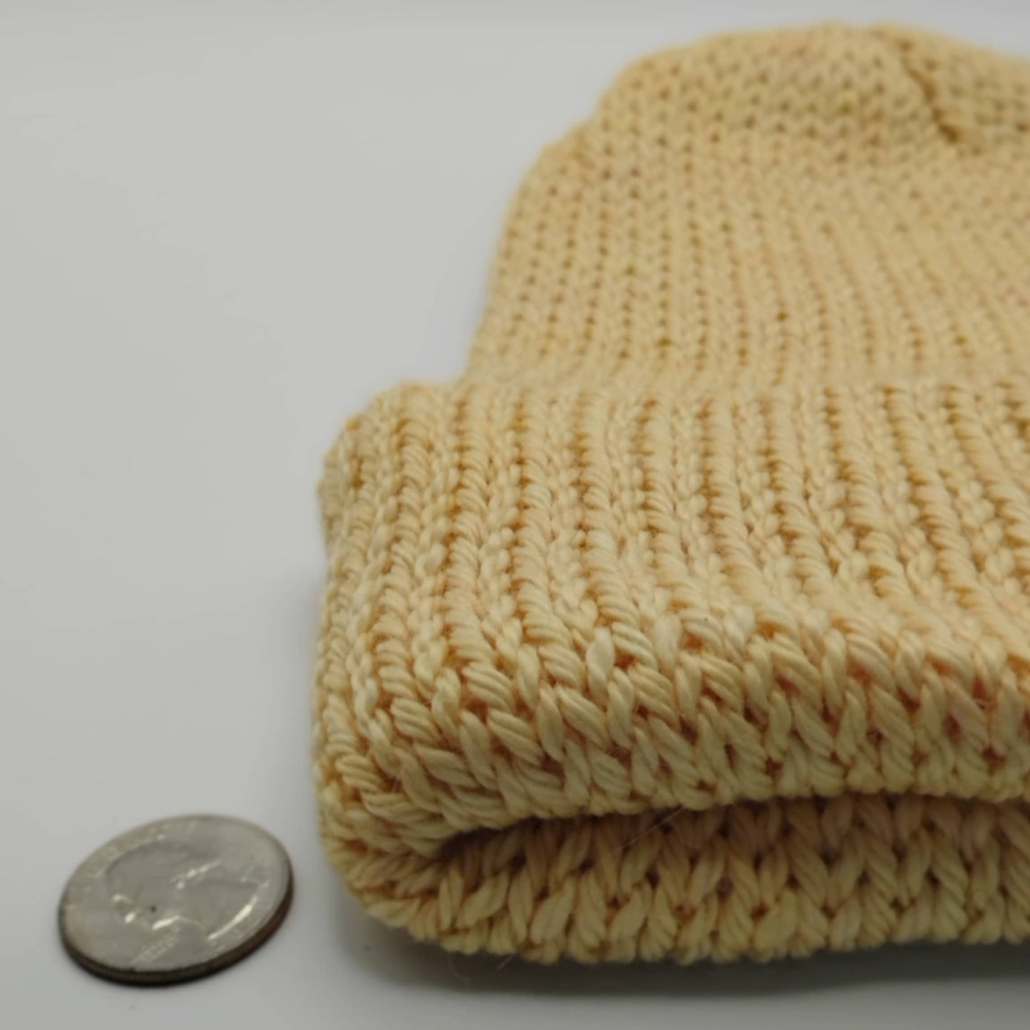 A close-up of a beige knit beanie, shown next to a coin for scale on a plain light background.