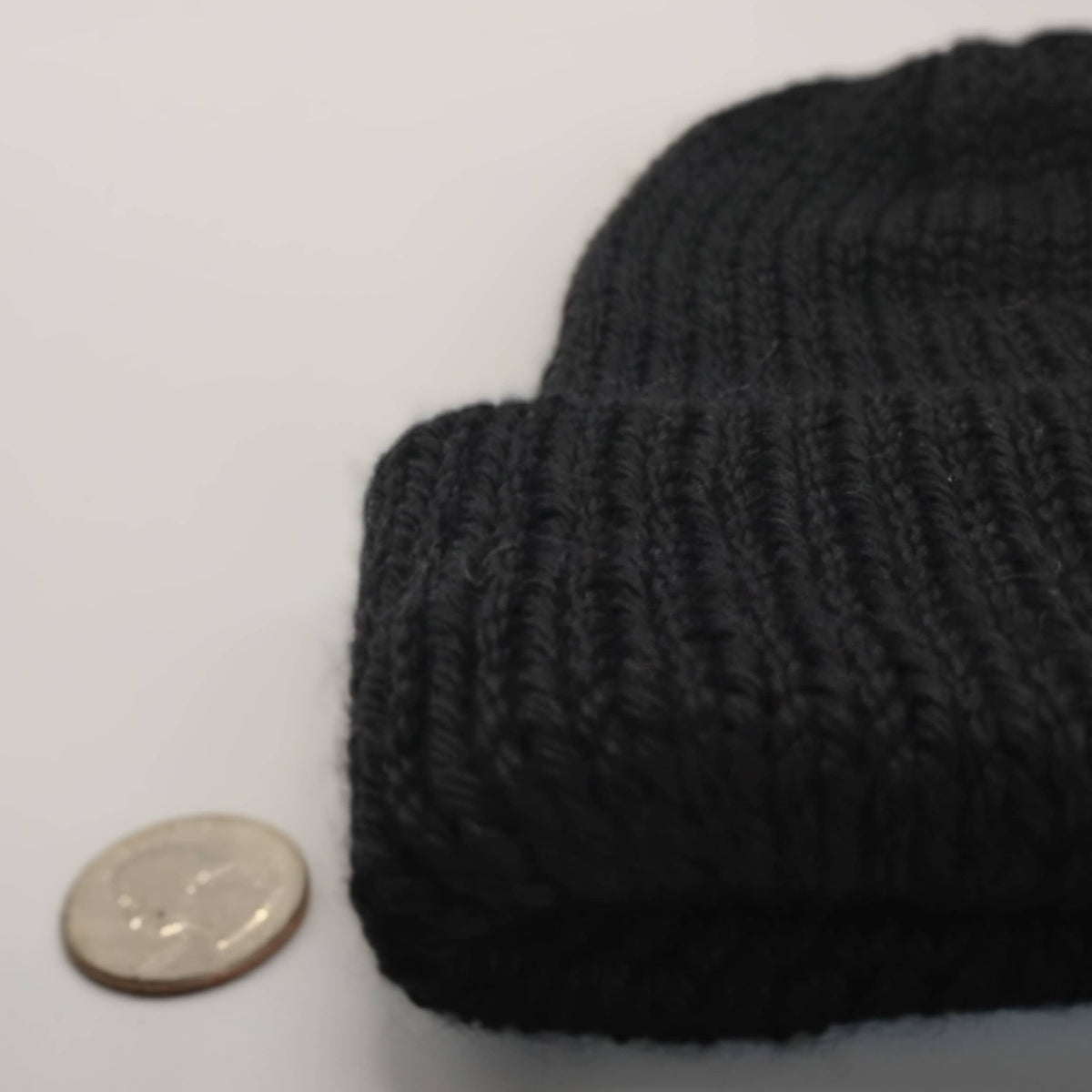 A close-up of a black knit beanie, shown next to a coin for scale on a plain light background.