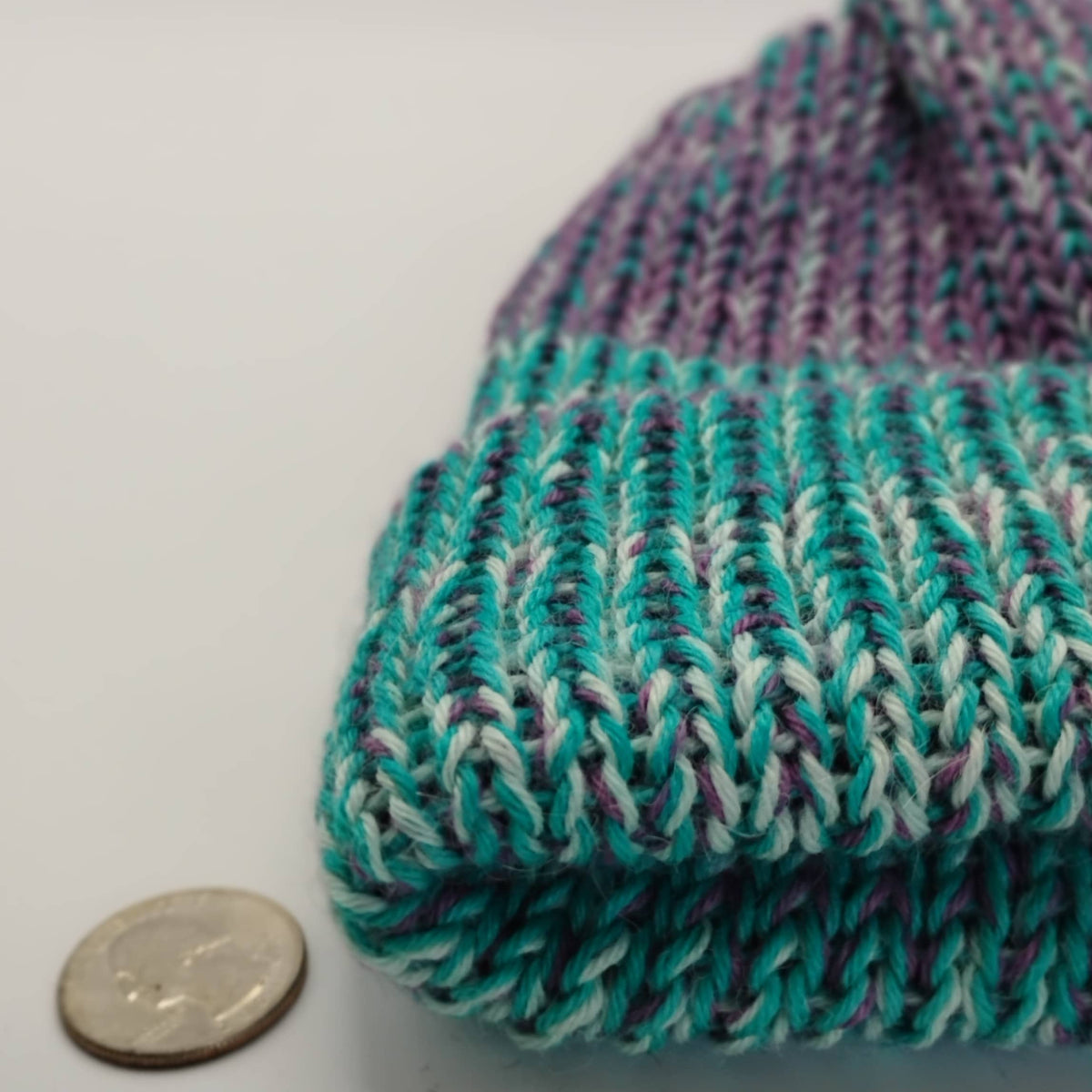 A close-up of a variegated blue knit beanie next to a US quarter coin for scale, on a plain light gray background.