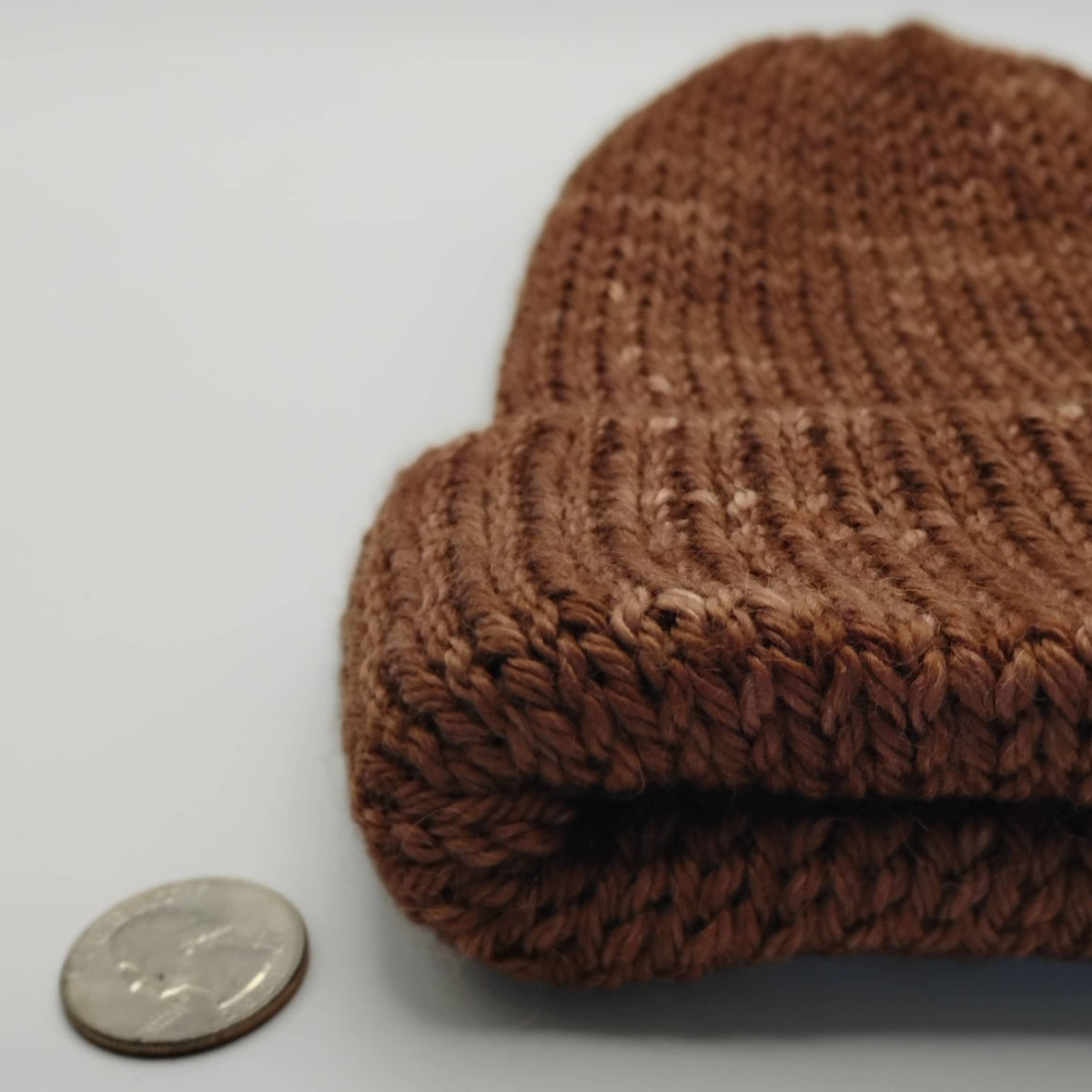 A close-up of a brown knit beanie, shown next to a coin for scale on a plain light background.