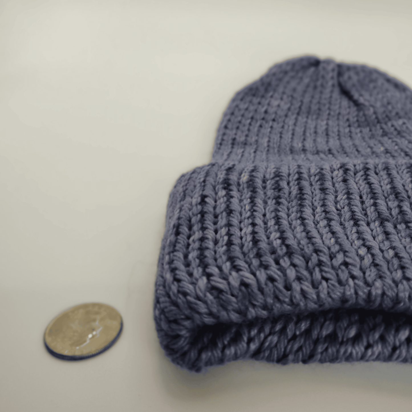 A close-up of a knit beanie in a denim color, shown next to a coin for scale on a plain light background.