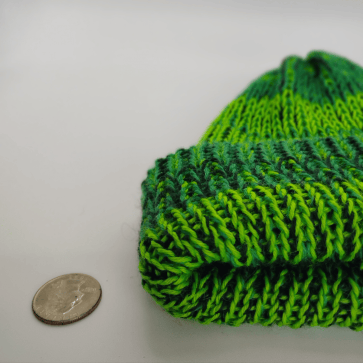 A close-up of a variegated green knit beanie next to a US quarter coin for scale, on a plain light gray background.