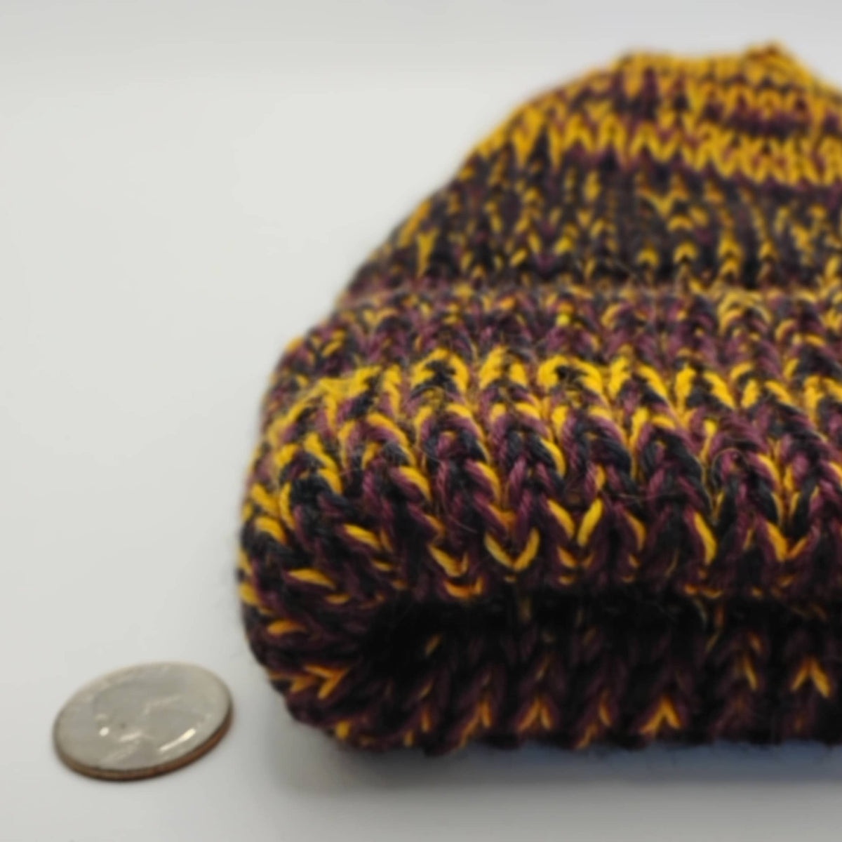 A close-up of a variegated maroon knit beanie next to a US quarter coin for scale, on a plain light gray background.