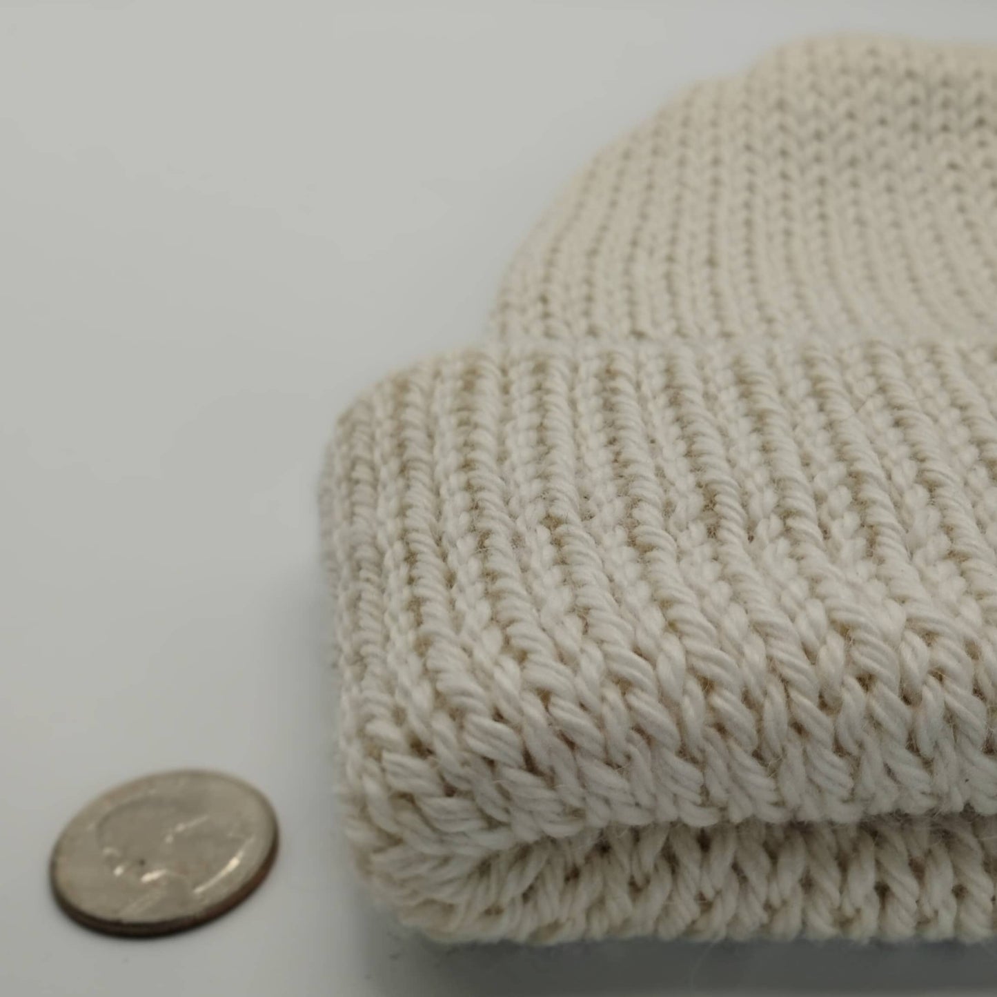 A close-up of a white knit beanie, shown next to a coin for scale on a plain light background.