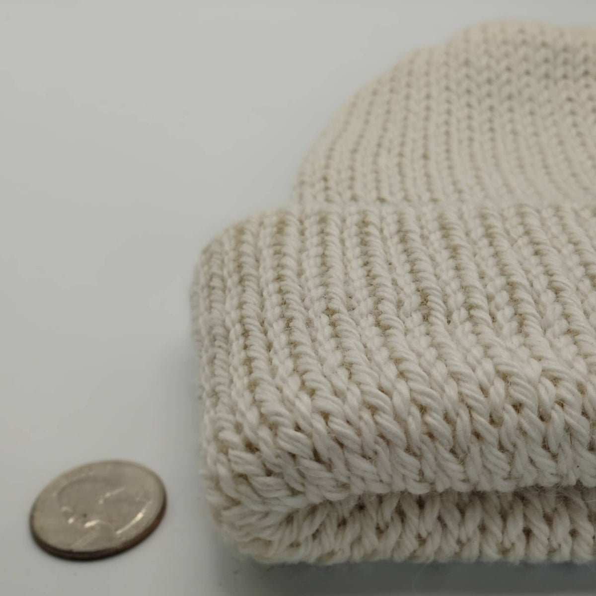 A close-up of a white knit beanie, shown next to a coin for scale on a plain light background.