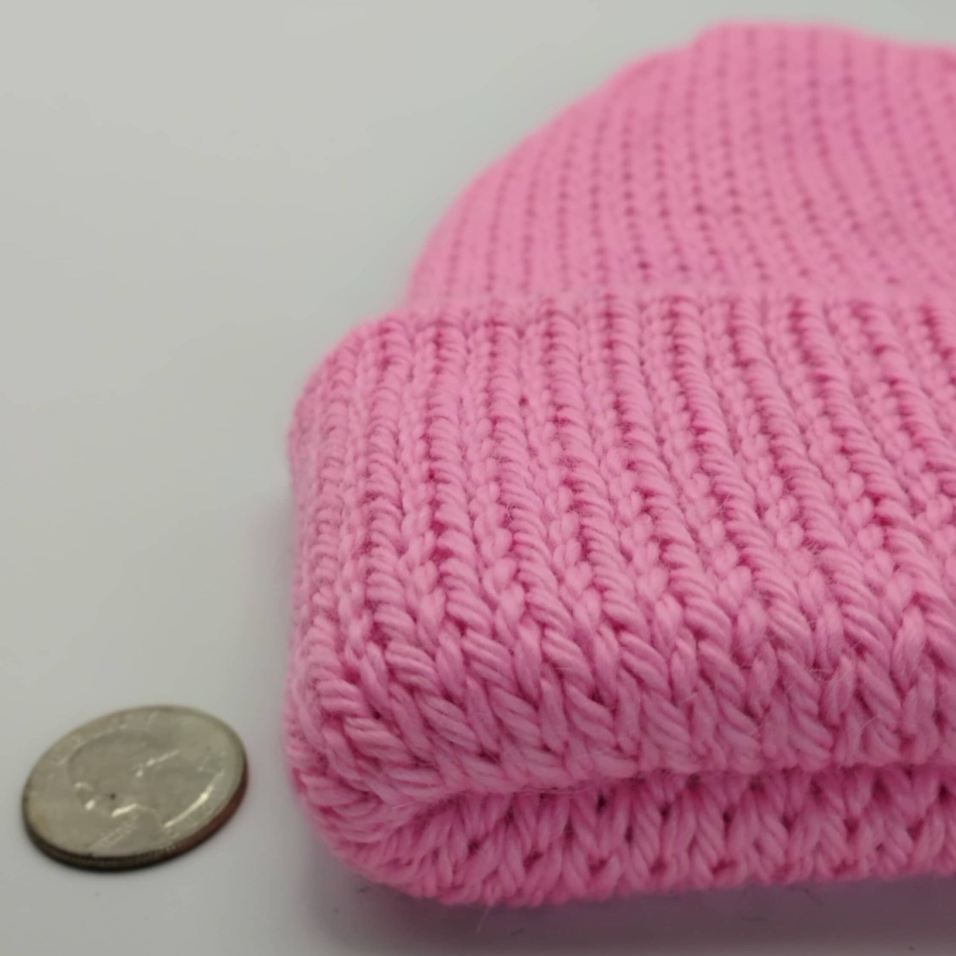 A close-up of a pink knit beanie, shown next to a coin for scale on a plain light background.