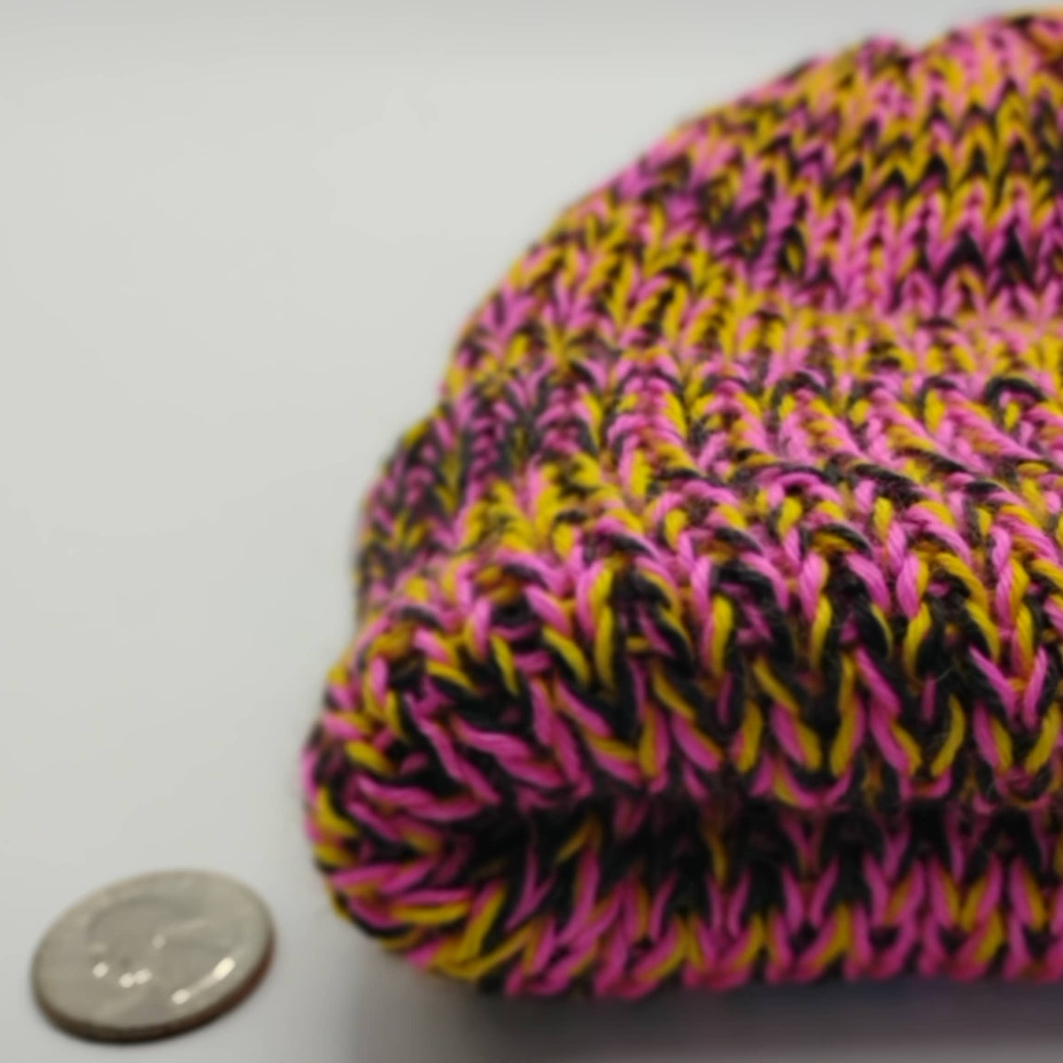 A close-up of a variegated pink knit beanie next to a US quarter coin for scale, on a plain light gray background.