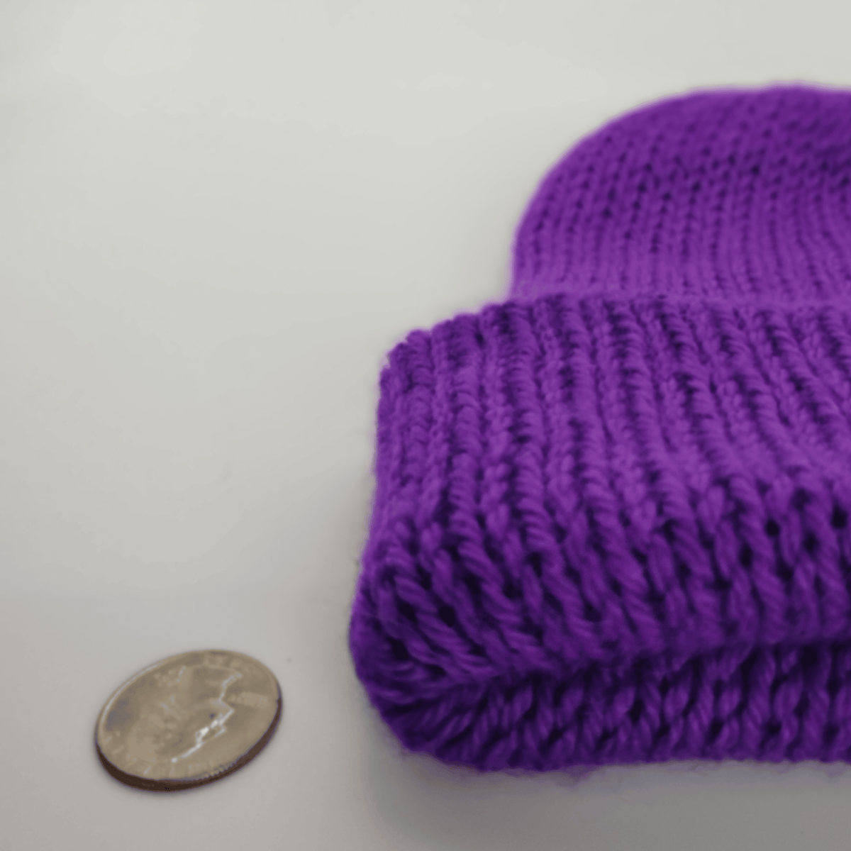 A close-up of a purple knit beanie, shown next to a coin for scale on a plain light background.