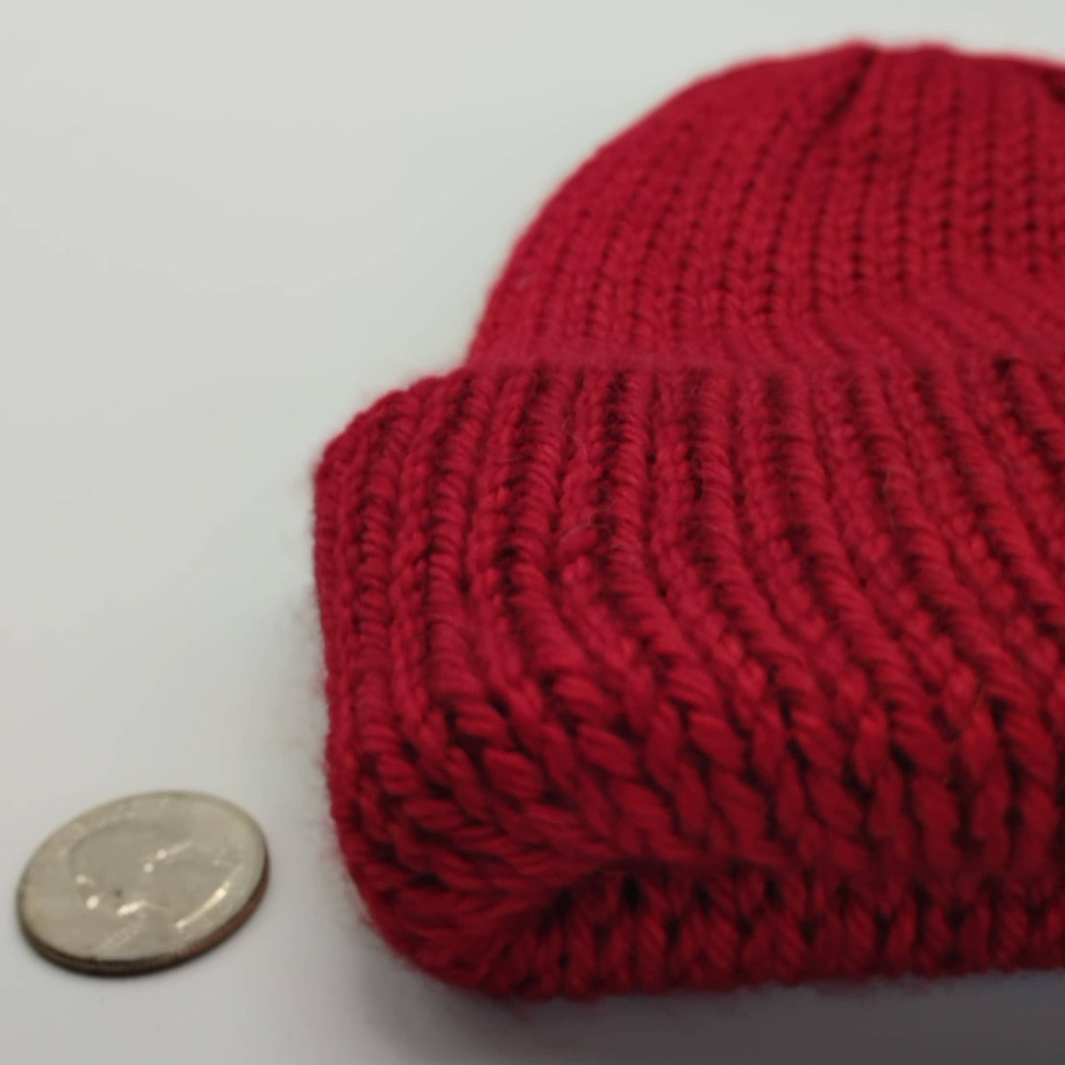 A close-up of a red knit beanie, shown next to a coin for scale on a plain light background.
