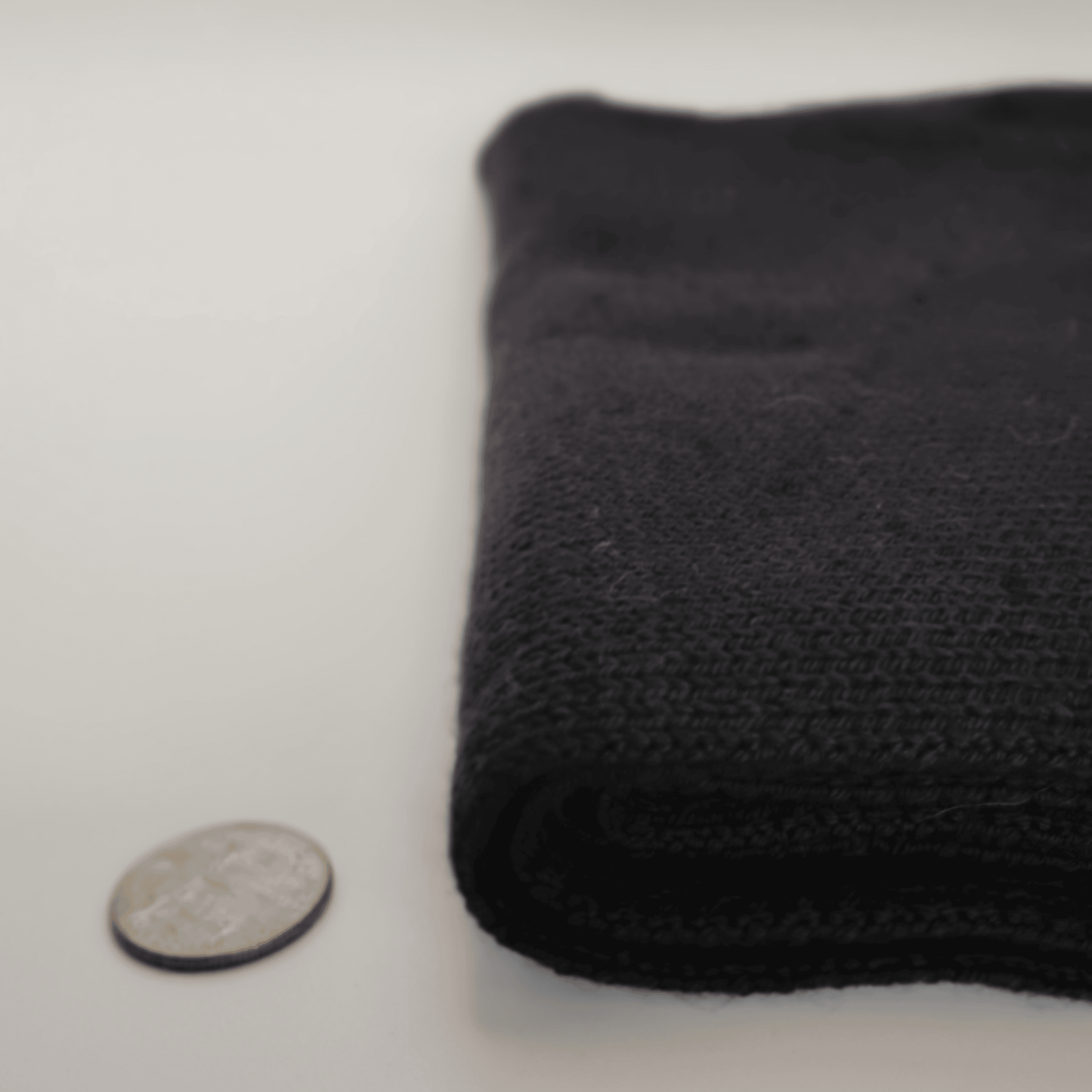 A folded black merino wool scarf shown on a white background next to a coin for scale.