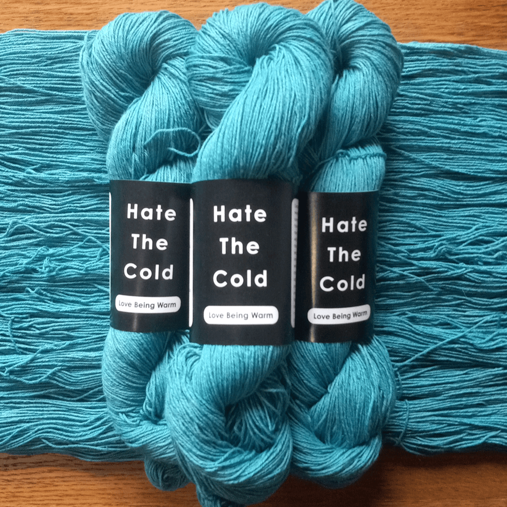 Three skeins of teal blue Cotton Linen yarn with black labels reading 'Hate The Cold' and 'Love Being Warm' laid on a wooden surface.