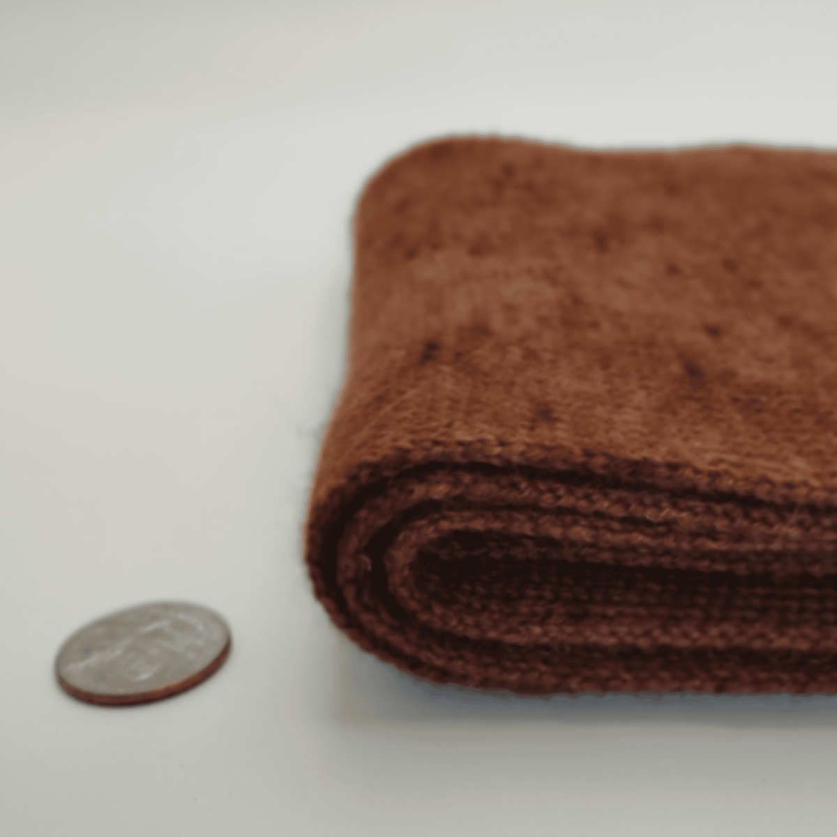 A folded brown alpaca scarf shown on a white background next to a coin for scale.