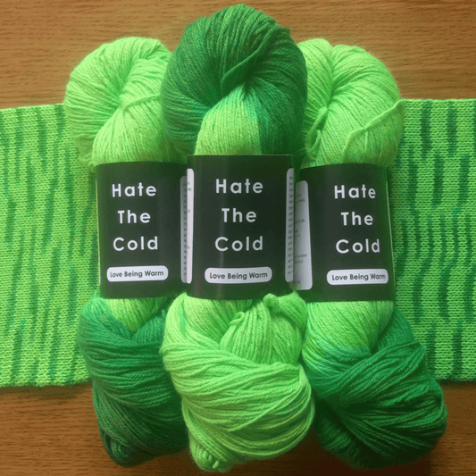 Three skeins of hankered yarn in bright green and lime green shades, arranged in front of a matching green scarf. Each skein has a black paper label with the text 'Hate The Cold' and 'Love Being Warm'.