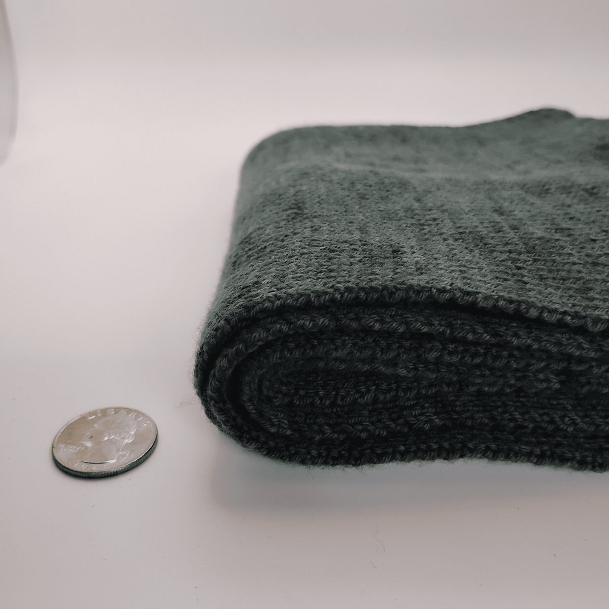 A folded dark green knit scarf, shown on a white studio background with a coin for scale.