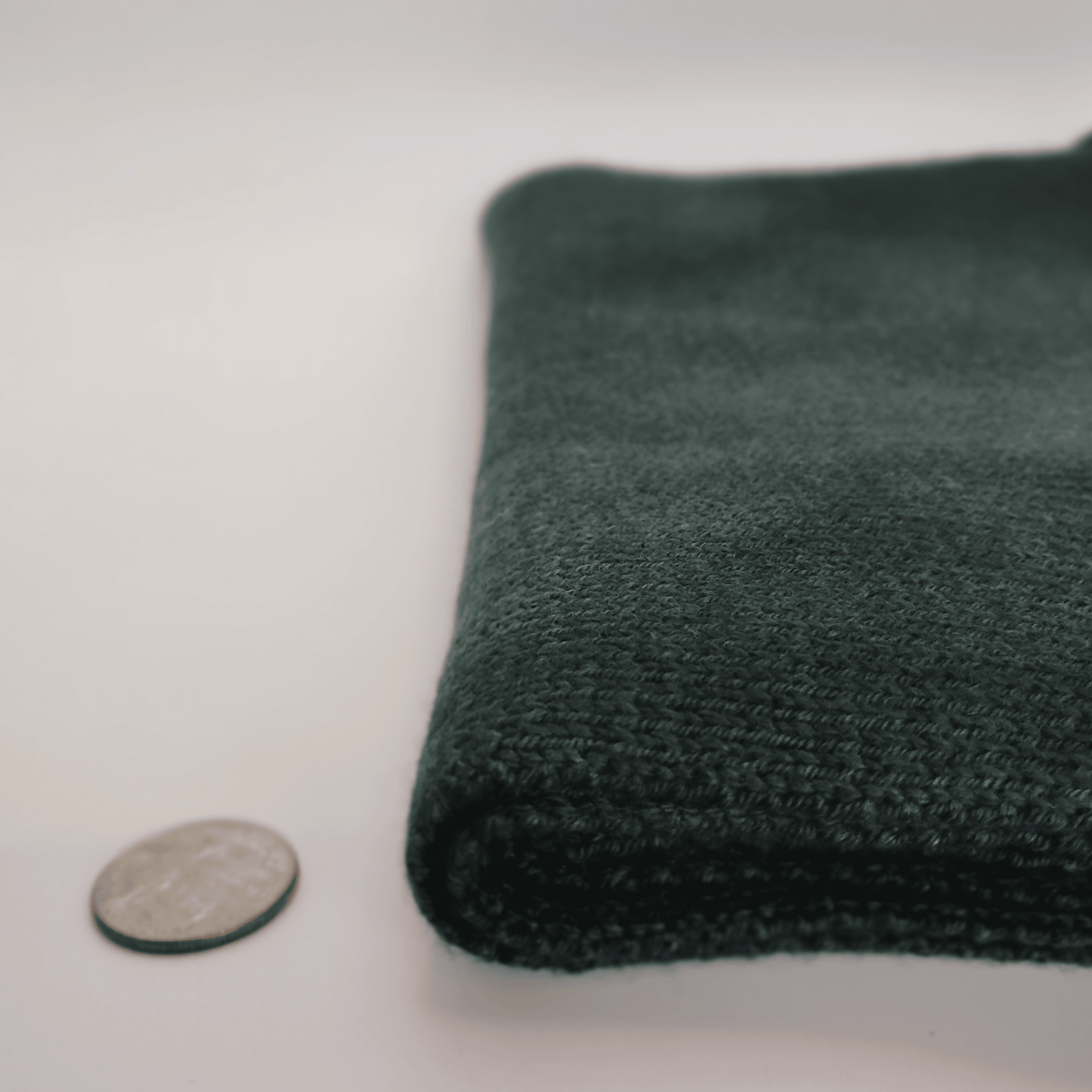 A folded dark green merino wool scarf shown on a white background next to a coin for scale.