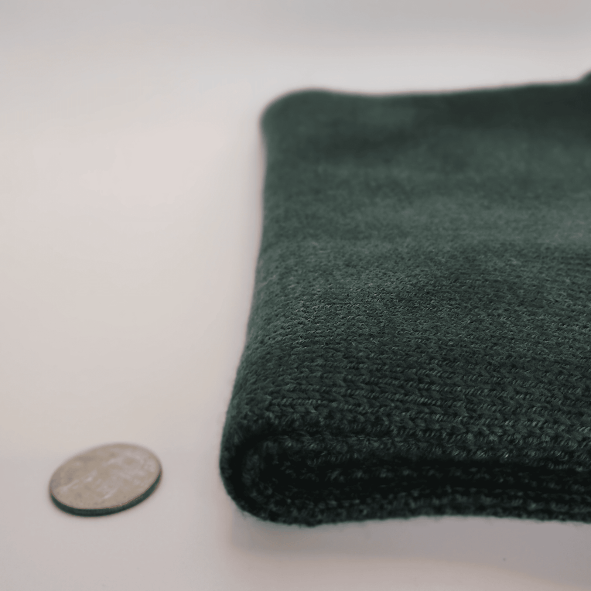 A folded dark green merino wool scarf shown on a white background next to a coin for scale.