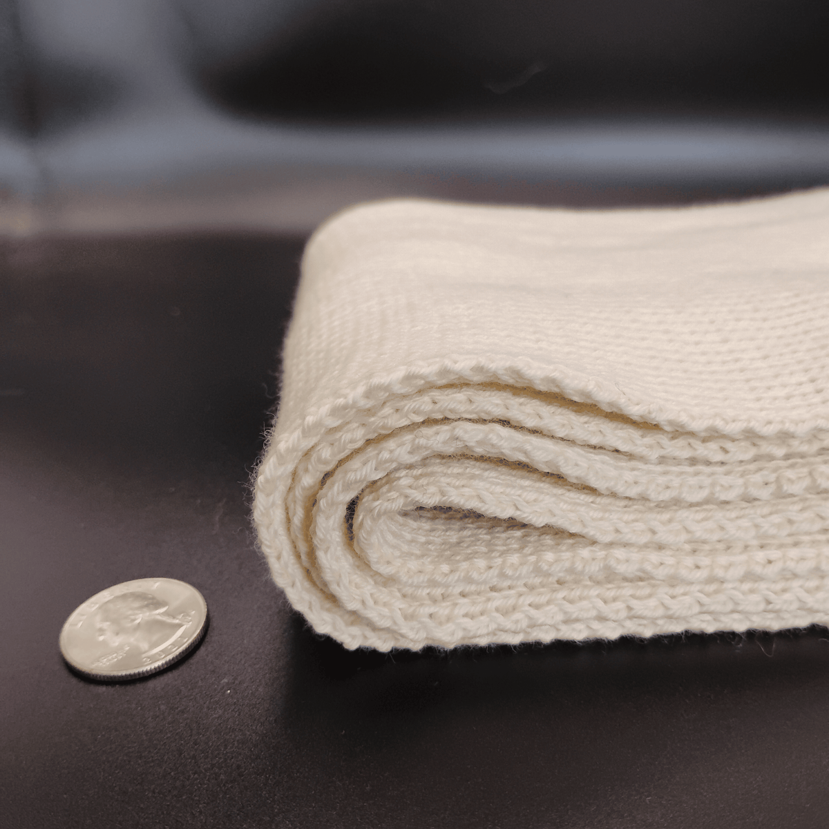 A close-up of a folded cream-colored knit scarf, shown next to a US quarter coin for scale on a dark surface.