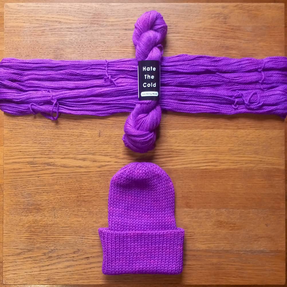 A purple knit beanie and matching purple yarn bundle laid flat on a wooden surface. The yarn bundle has a black paper label that reads 'Hate the Cold'.