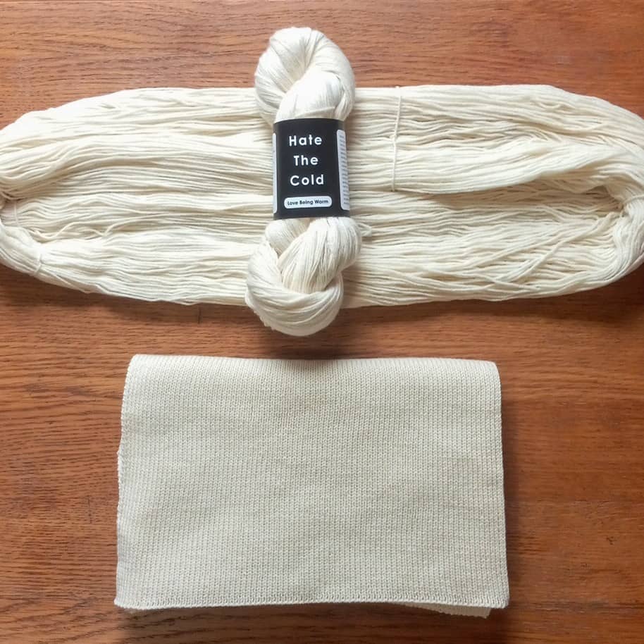 Natural white merino wool yarn and matching knit scarf laid flat on a wooden surface, with a black label reading 'Hate The Cold' on the yarn.