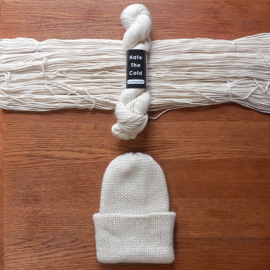A knitted beanie in natural white displayed below a matching skein of yarn on a wooden surface. The yarn has a black paper label wrapped around it that reads 'Hate The Cold'.